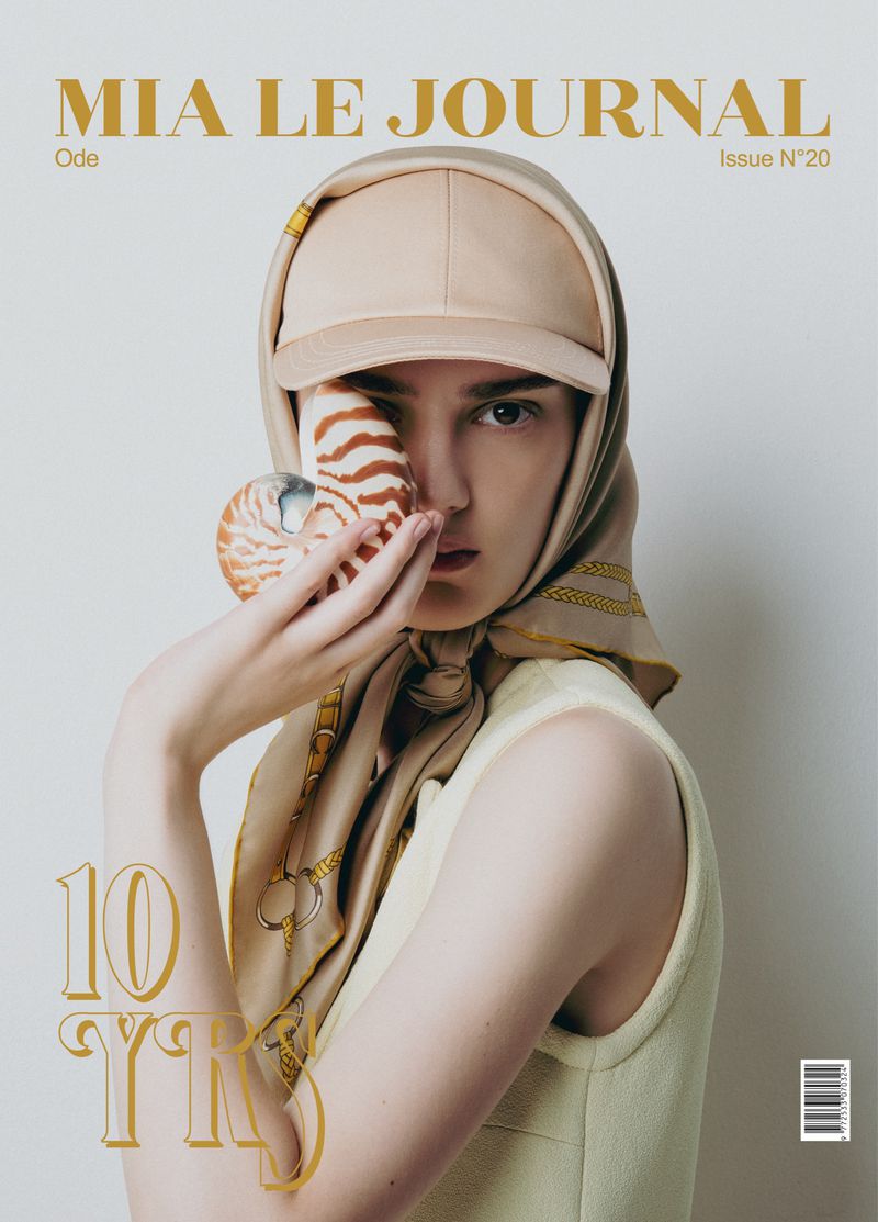 Fashion magazine cover featuring a woman in a headscarf and baseball cap holding a nautilus shell to her face in a studio.