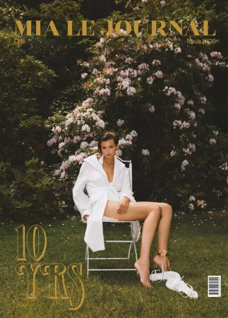 A woman in a white belted robe sits on a white folding chair outdoors on a lawn before blooming flower bushes.