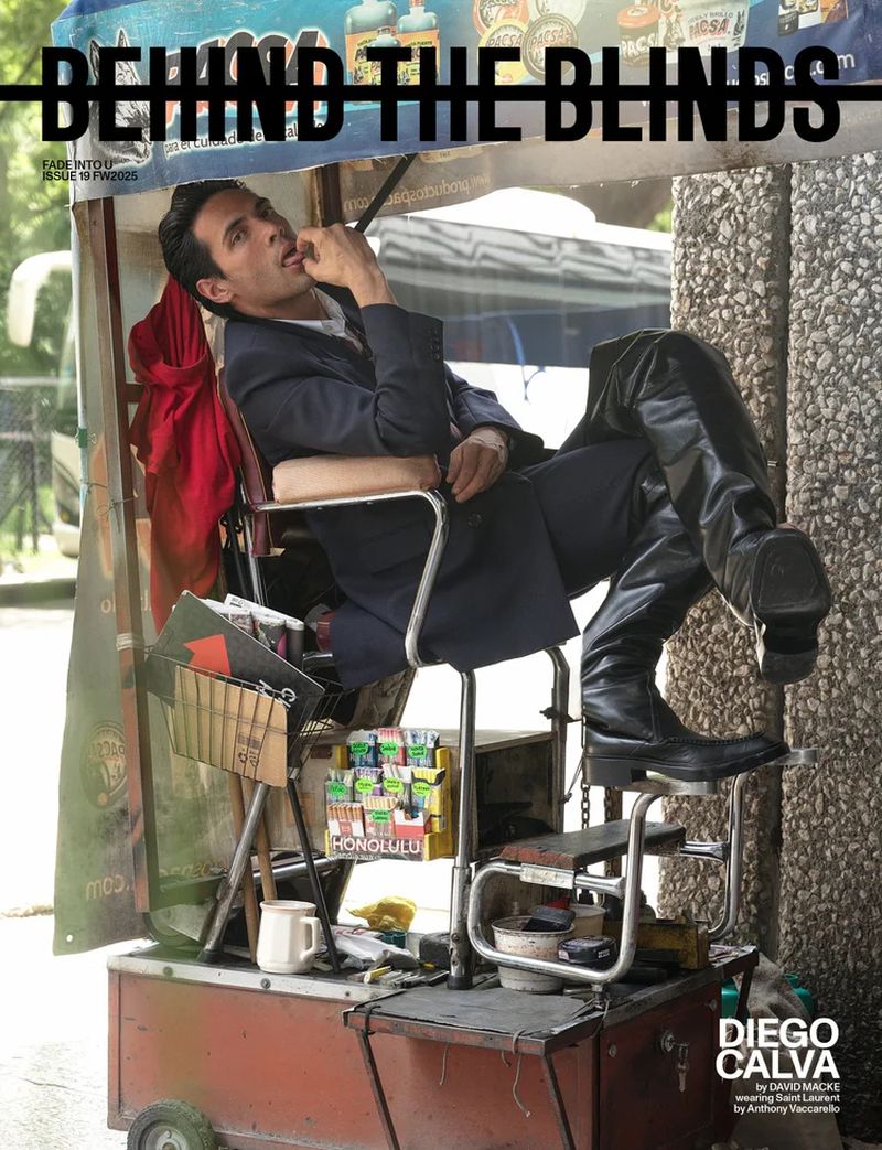 Diego Calva reclines on a shoe-shine cart, striking a pose in a suit and boots, with a magazine cover behind him in an urban setting.
