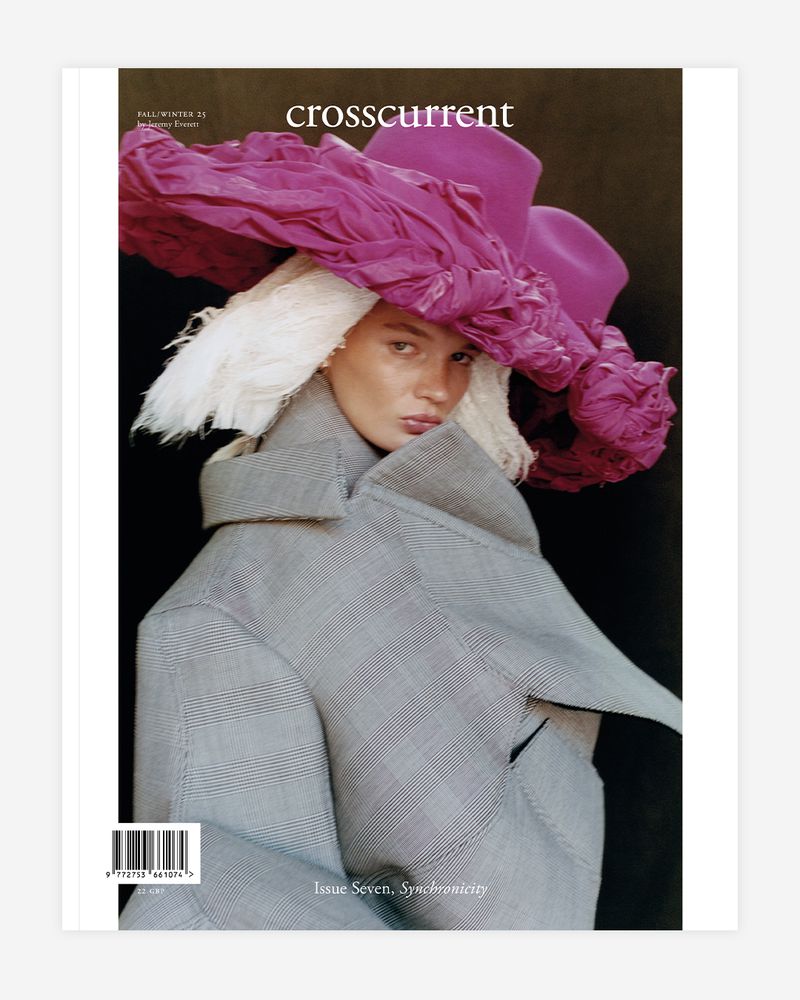 Fashion editorial featuring a woman with white hair wearing a pink hat and grey coat in a studio setting. Magazine cover.