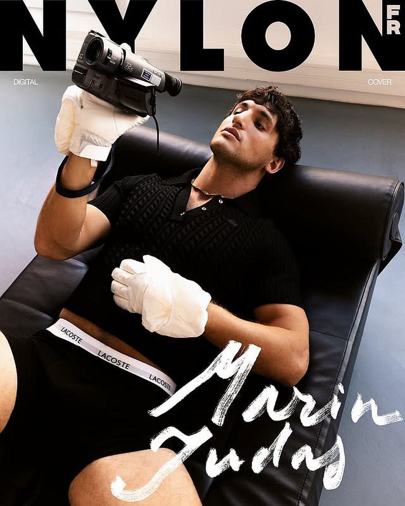 Man lying on black chaise lounge chair with a vintage video camera and white gloves, against a studio backdrop with magazine cover text.