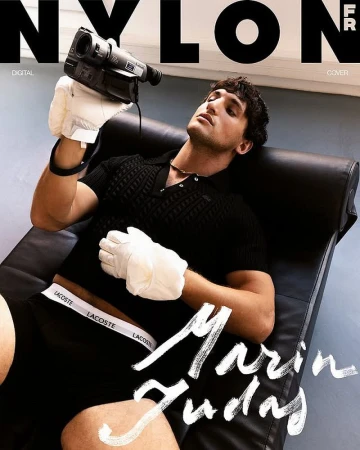 Man lying on black chaise lounge chair with a vintage video camera and white gloves, against a studio backdrop with magazine cover text.