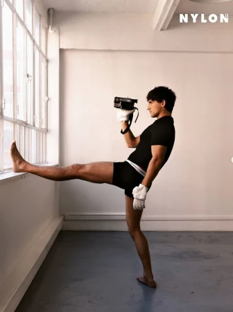 Asian man in athletic wear poses indoors with a vintage camcorder, leg raised against a window, with the NYLON logo in the corner.