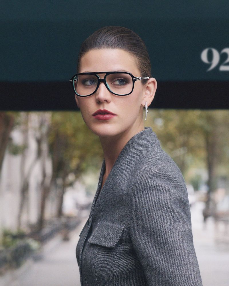 Attractive woman poses wearing aviator eyeglasses and a gray tweed jacket against an urban background with natural lighting.