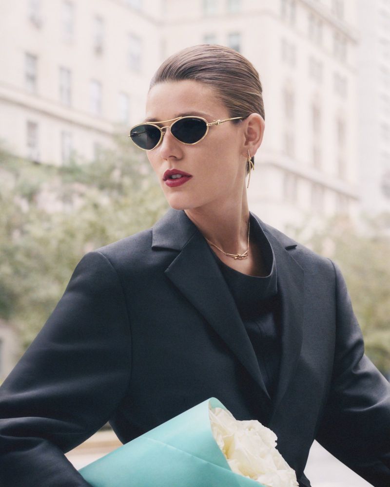 Woman in sunglasses and black blazer poses outdoors holding bouquet. Close-up fashion shot with city buildings in the background.