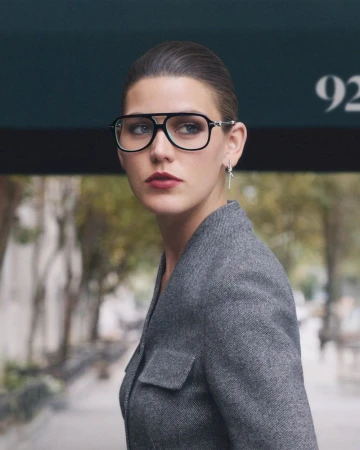 Model Vivienne Rohner wears black aviator eyeglasses and a tailored grey jacket beneath a dark awning on a city street.