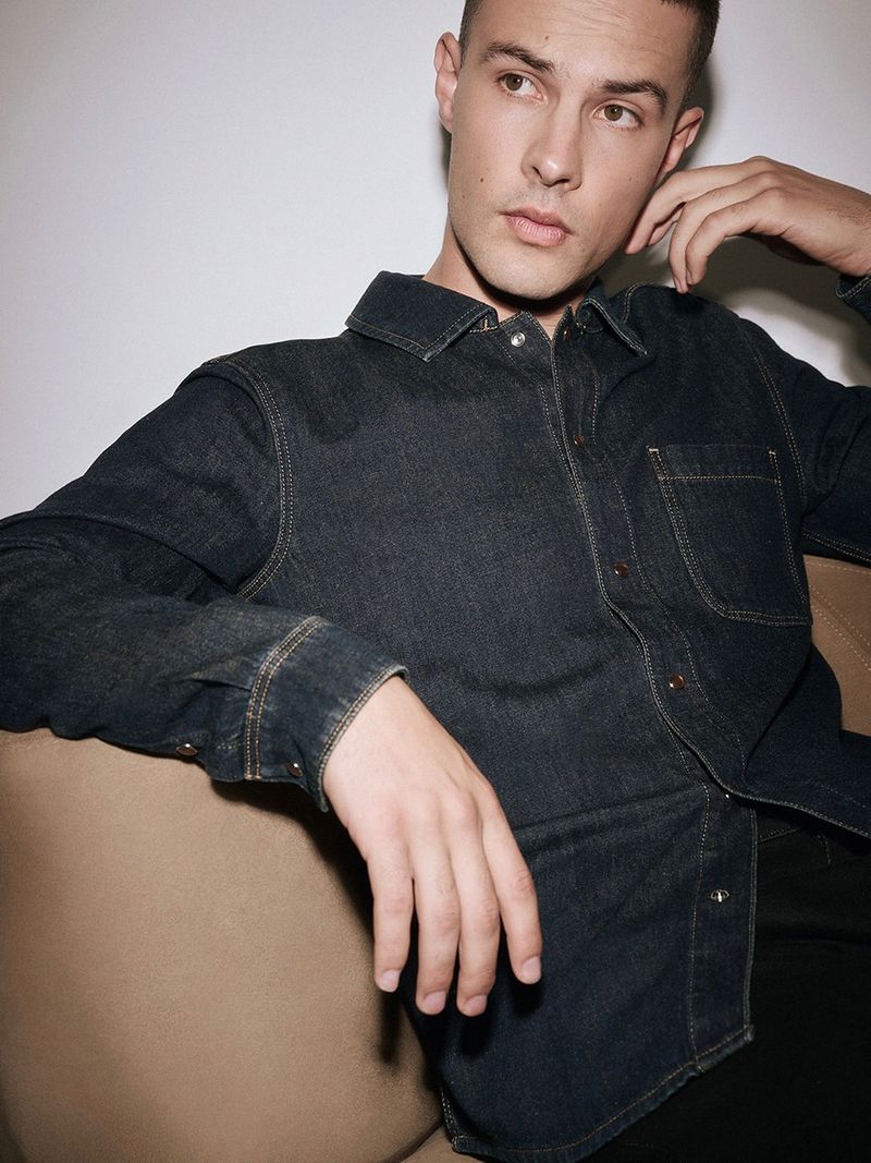 Young adult male in a dark denim shirt leaning on a tan couch in a casual indoor setting with soft, diffused lighting.