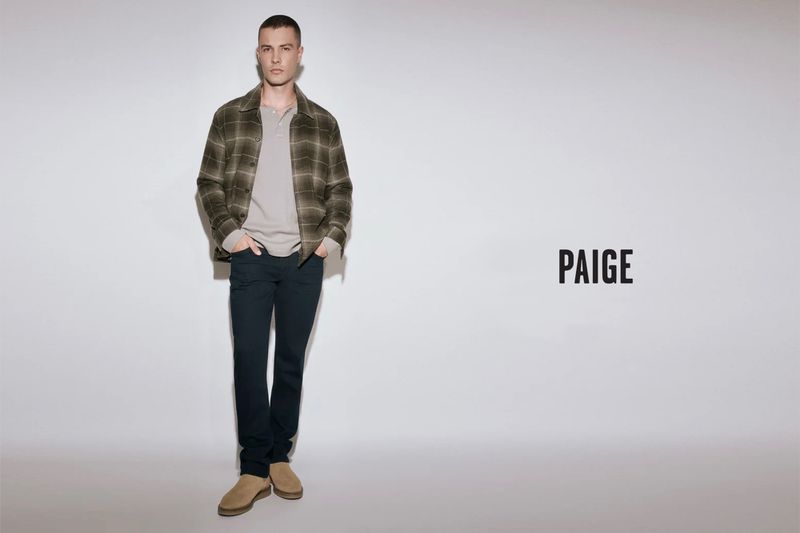 A man in a light shirt, plaid jacket, dark pants, and tan shoes poses against a white wall with the brand PAIGE to the right.