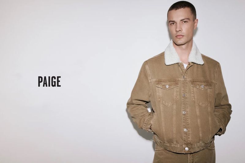 A fair-skinned adult male model poses in a studio, wearing a brown denim jacket with a white sherpa collar, paired with matching pants.
