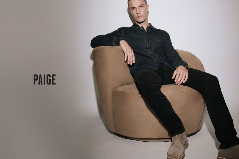 A fair-skinned adult male lounges in a tan armchair wearing a dark denim shirt, black jeans and tan suede shoes against a grey background.