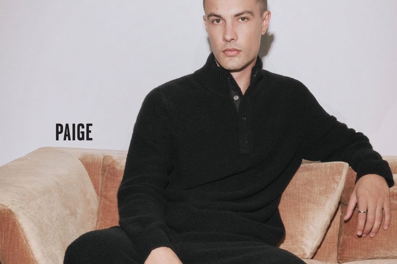 A male model poses on a light brown sofa indoors. The man is wearing a dark sweater. The photo is a product shot for PAIGE.