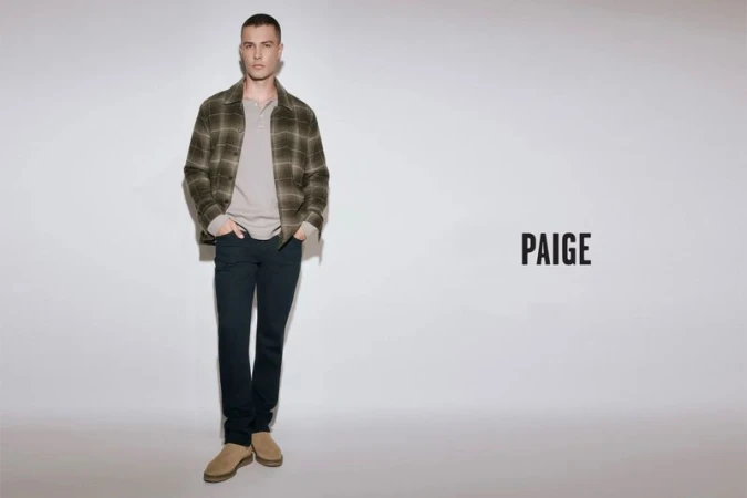 Full-length portrait of a male model wearing a plaid jacket, light grey henley, dark jeans, and tan boots against a plain grey studio backdrop