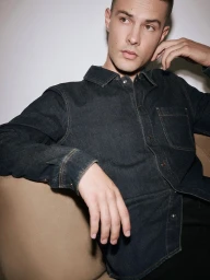 Close-up portrait of a male model wearing a dark denim shirt, seated indoors against a plain wall.