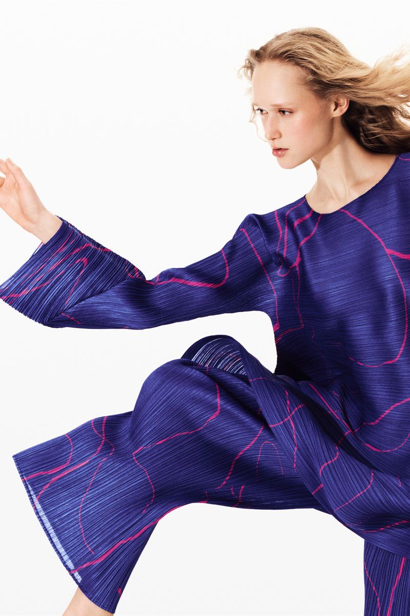 Woman wearing a blue pleated long-sleeve top and pants with a pink wavy pattern, captured in a high-key fashion shot with a white background.