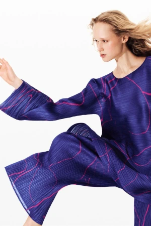 Woman wearing a blue pleated long-sleeve top and pants with a pink wavy pattern, captured in a high-key fashion shot with a white background.