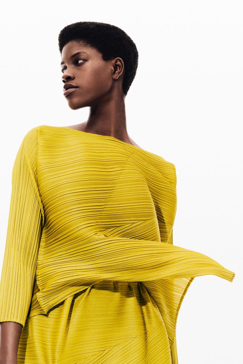A Black woman wearing a pleated chartreuse dress looks over her shoulder in a studio portrait against a white background. Fashion shot.