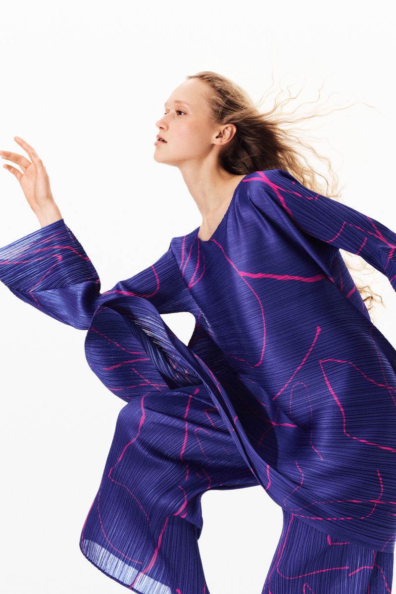 Blonde woman models a blue pleated pant suit with pink pattern in a bright, high-key fashion studio shot with dramatic pose.