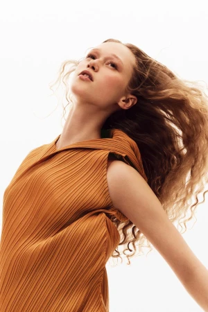 Portrait of a woman with curly blonde hair wearing an orange pleated top looking up against a bright white background in a fashion shot.
