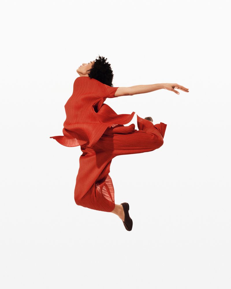 A woman in motion, jumping with outstretched arms and legs, wearing a pleated orange outfit against a stark white backdrop in a studio setting.