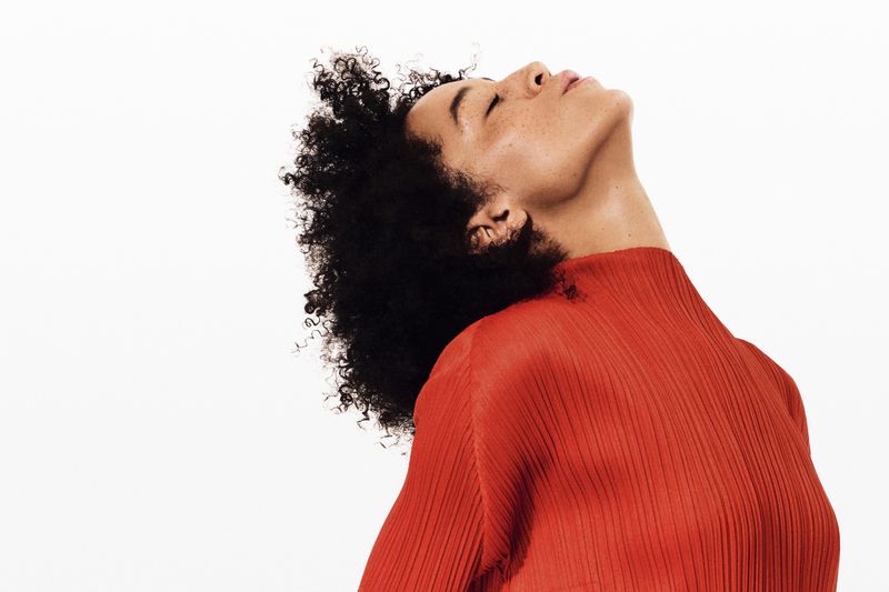 Portrait of a Black woman with an afro tilting her head back, wearing a ribbed orange top, against a white background.