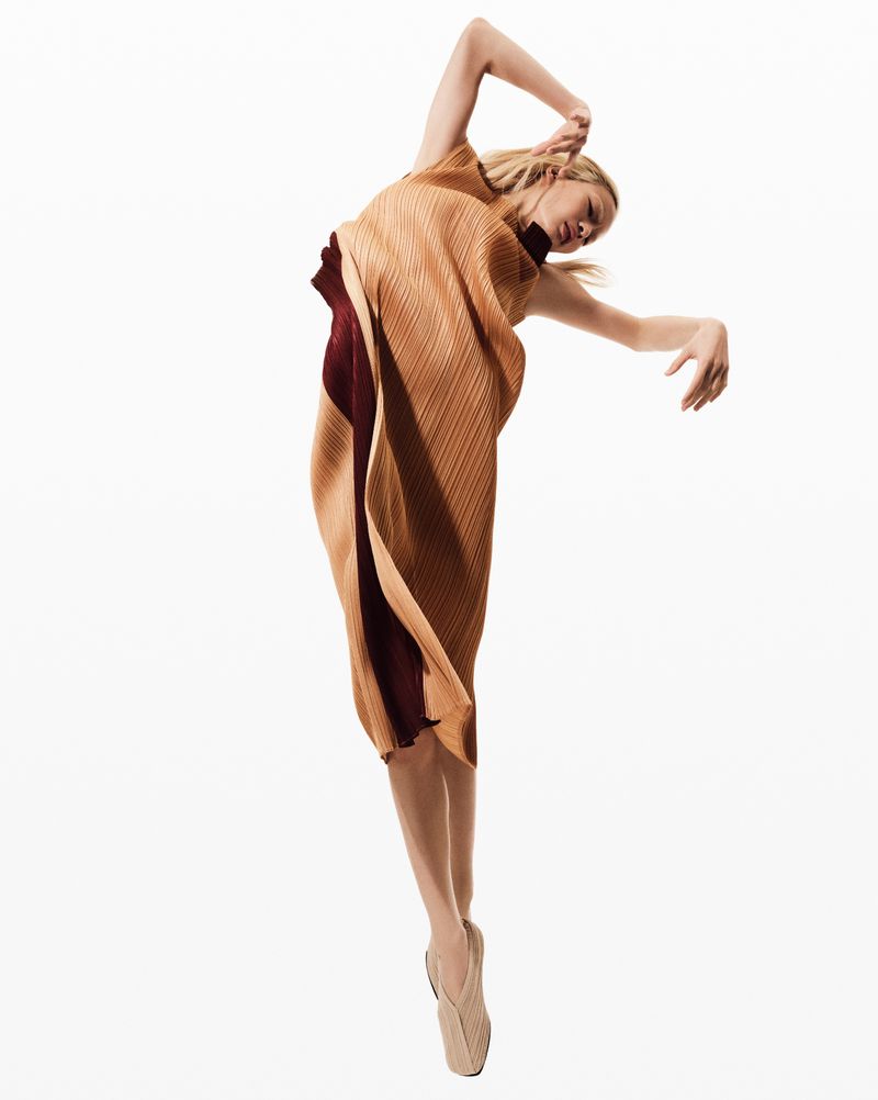 Elegant woman in flowing brown dress poses gracefully in a studio setting, arms raised, legs crossed, and wearing ballet-style shoes.