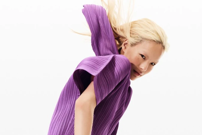 Blonde haired woman wearing a pleated purple top in a fashion headshot on a white background in a brightly lit photo studio.