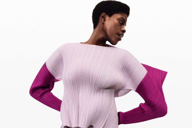 Fashion shot of an African-American woman in a lilac, plissé top with magenta sleeves, posing in a photo studio with a white background.
