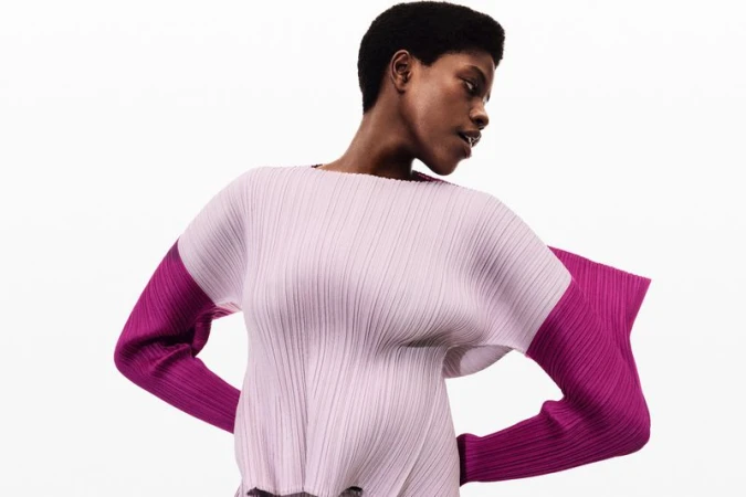 Fashion shot of an African-American woman in a lilac, plissé top with magenta sleeves, posing in a photo studio with a white background.