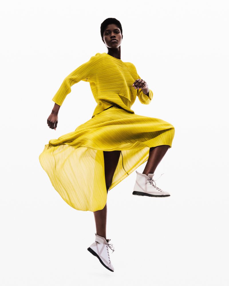 Fashion shot of a woman jumping, wearing a yellow outfit and white sneakers, set against a clean white background in a studio setting.