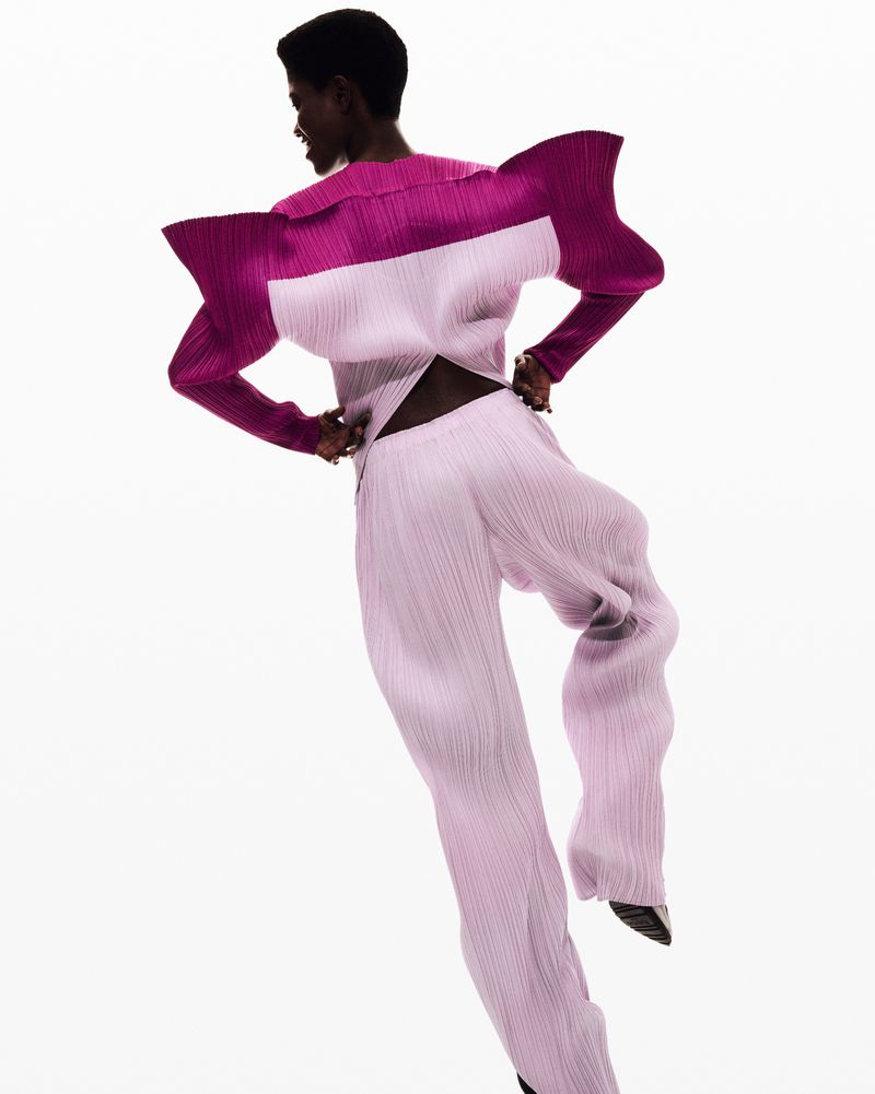 Fashion model strikes a pose in a magenta and lavender pleated outfit against a bright white backdrop in a studio setting.