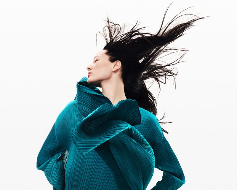 A fashion portrait features a woman in a textured, teal, flowing top against a white background, her dark hair catching the light.