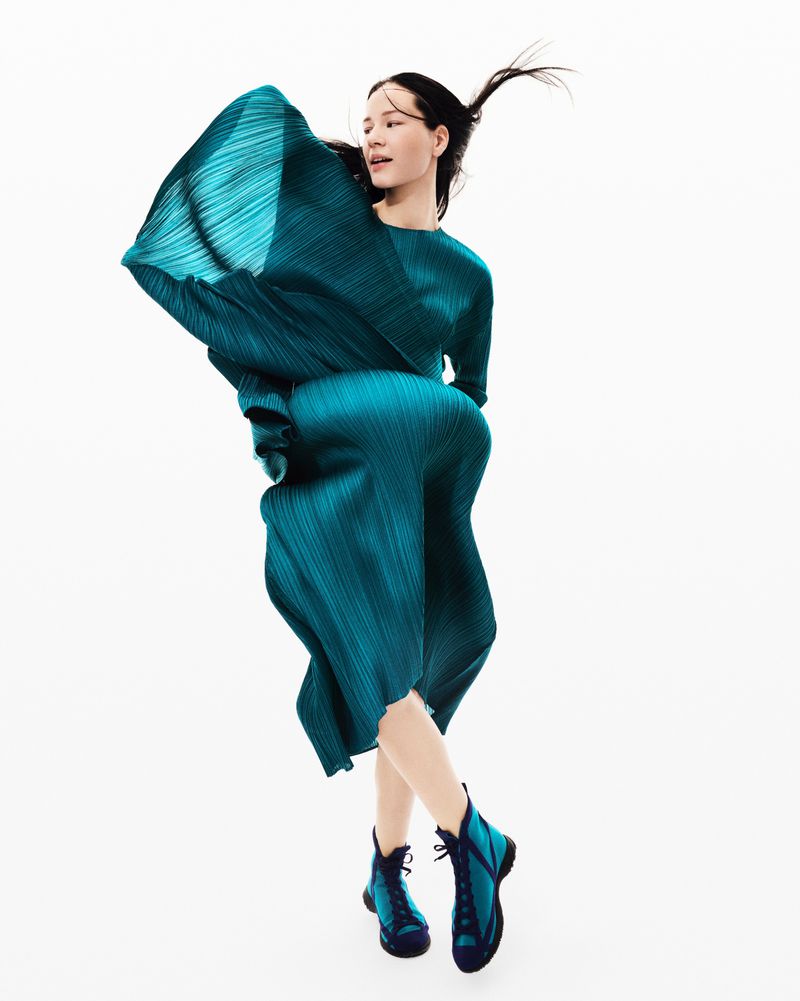 Woman in a dynamic pose, wearing a blue pleated dress and boots against a bright background, showcasing fashion and style.