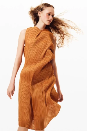 Model with windblown wavy hair wearing a sculptural pleated mustard brown sleeveless dress against a white studio background.