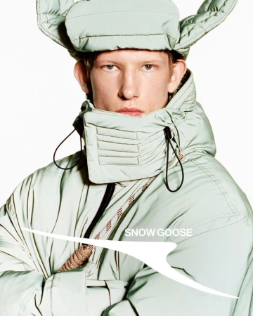 Fashion shot of a person wearing a winter hat and a light green, puffy, insulated coat against a stark white background.
