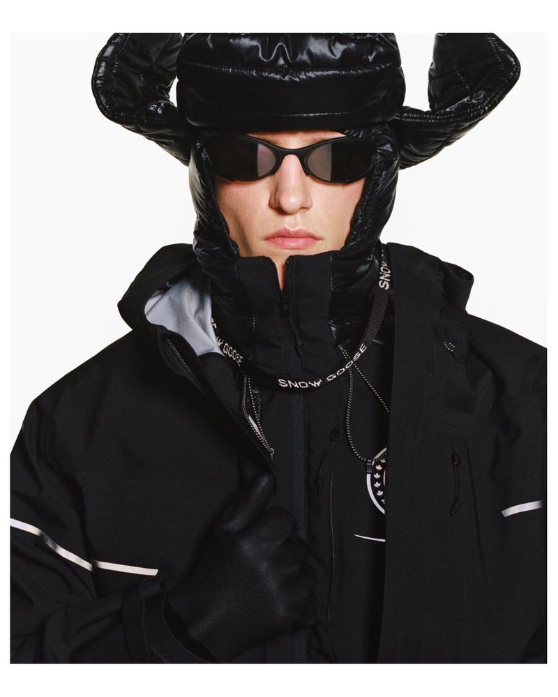 Fashionable adult male wearing black winter coat with fur-lined hood, black hat, sunglasses, and gloves, in a studio shot.