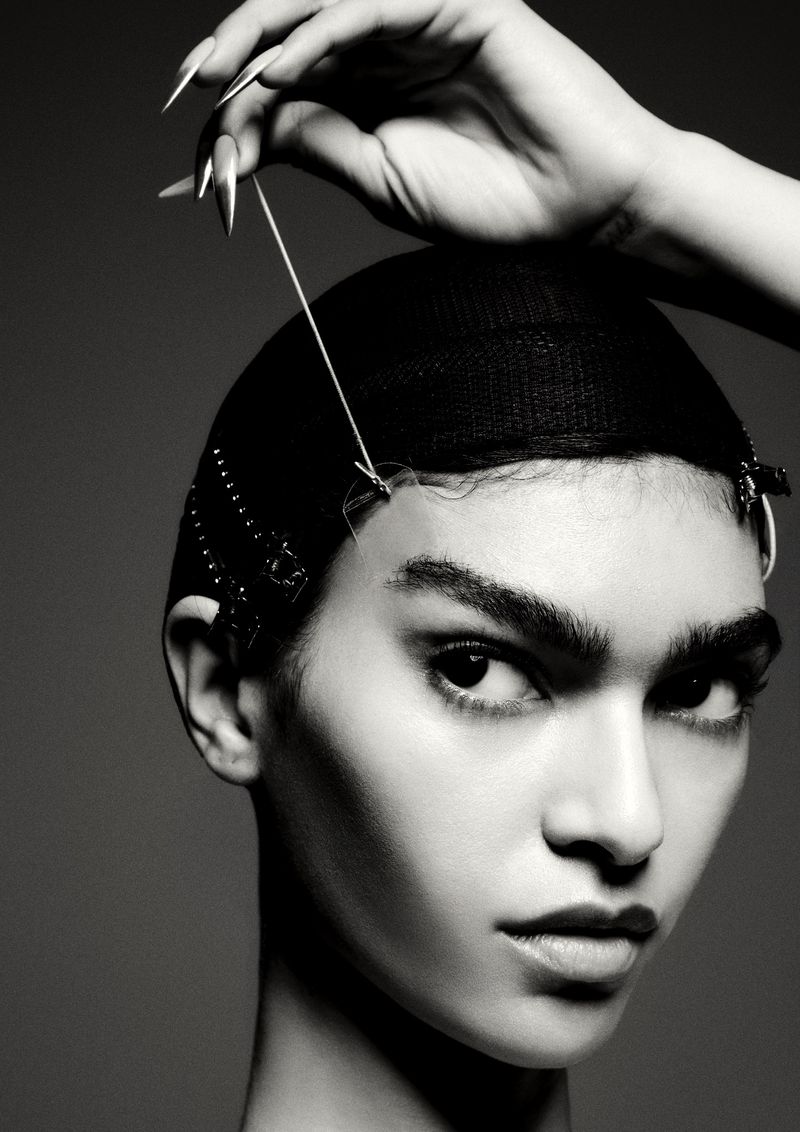 Striking black and white portrait of a woman with a net cap, a hand holding a thread to the cap, and metal clips along the hairline.