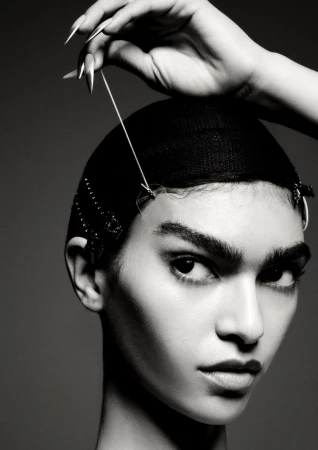 Striking black and white portrait of a woman with a net cap, a hand holding a thread to the cap, and metal clips along the hairline.
