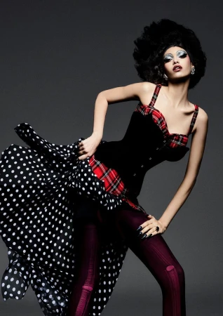 Elegant woman in a black bustier, plaid accents, and a polka dot skirt, posing in a studio with dark hair and striking makeup.