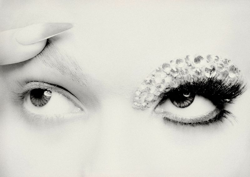 Close-up black and white image of a face with one eye showing decorative rhinestones on the eyebrow and eyelash with a hand.