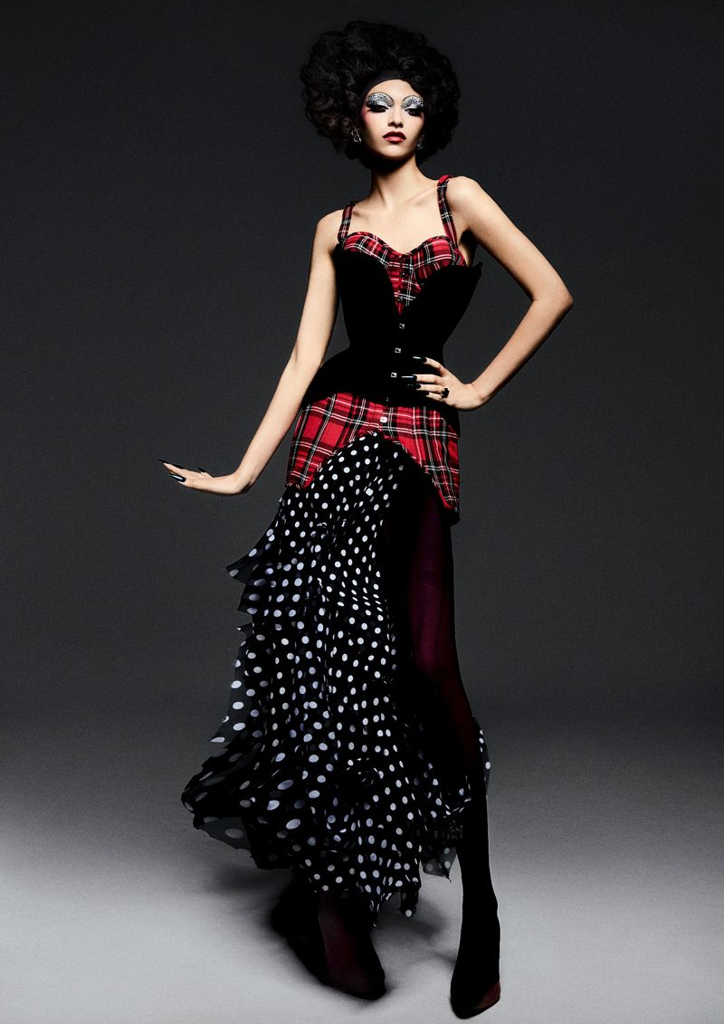 A slender fashion model in an avant-garde red plaid bodice, black velvet corset, and black and white polka dot skirt poses dramatically.