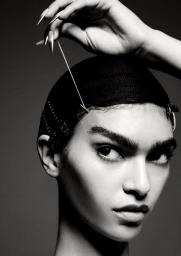 Close-up monochrome portrait of a model with long metallic fingernails holding a thread against a dark background.