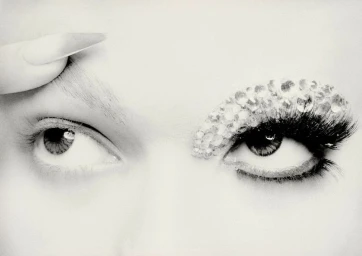 Extreme close-up of a model's eyes in black and white, featuring one bare eye and one embellished with crystal rhinestones and false lashes.