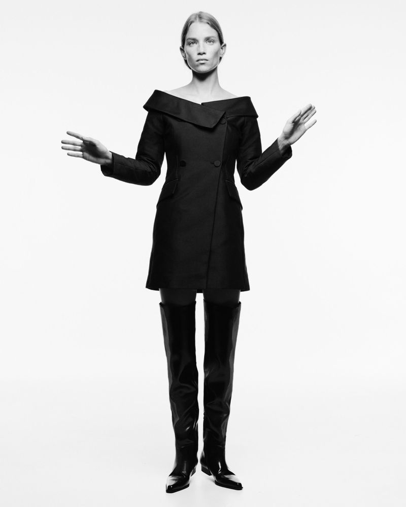 A fashion image in black and white features a poised, adult female model wearing a modern off-the-shoulder jacket dress and thigh-high boots.