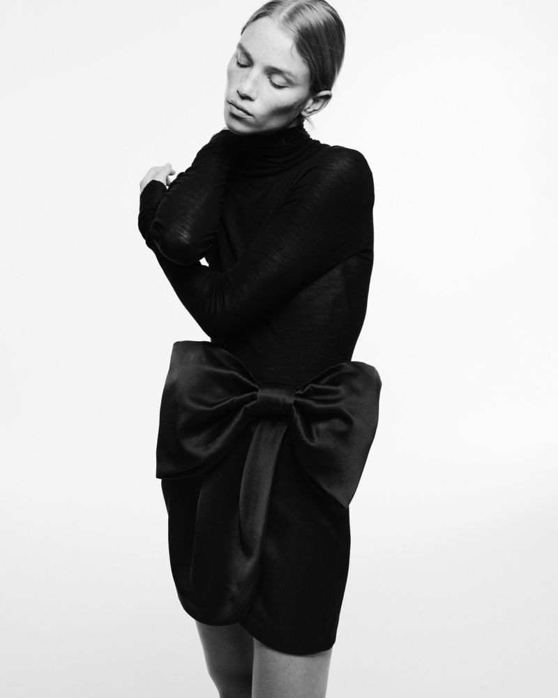 Monochrome studio shot of a woman in a black dress with a large satin bow. She has closed eyes and crossed arms.