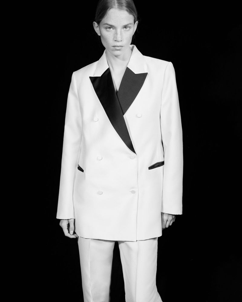Studio shot of a woman in a white suit with black lapels, standing against a black backdrop in a fashion-editorial style.