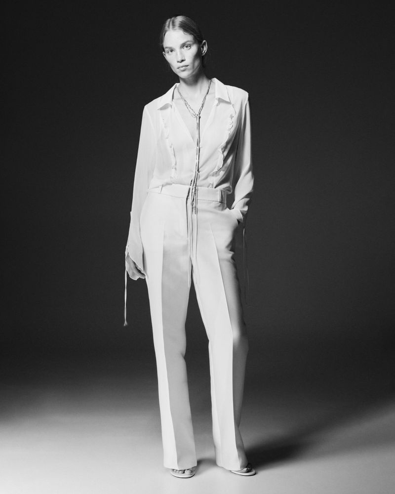 Black and white studio shot of a female fashion model posing in a white blouse and pants, wearing metallic heels and a rope necklace.