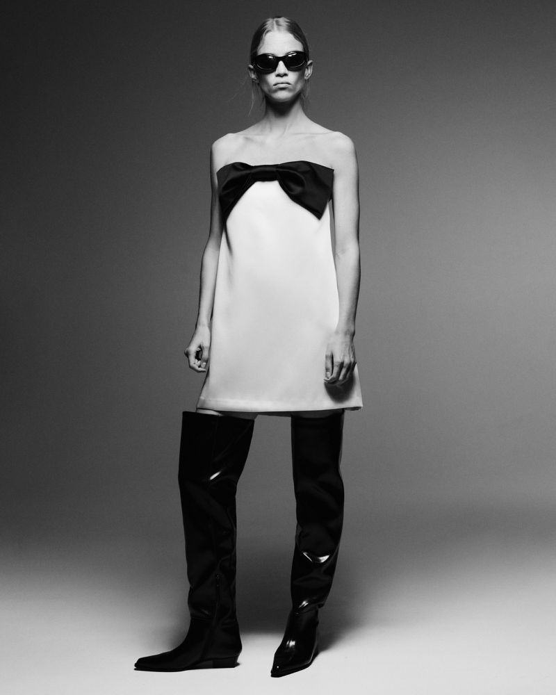 Rebecca Leigh Longendyke wearing a white strapless mini dress with a black bow and thigh-high black boots against a studio backdrop.