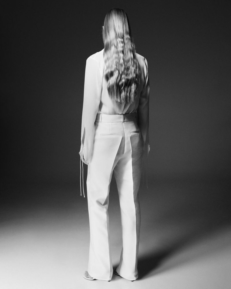 Back view of a woman with long wavy hair wearing a white blouse and flared trousers in a studio with a soft light.