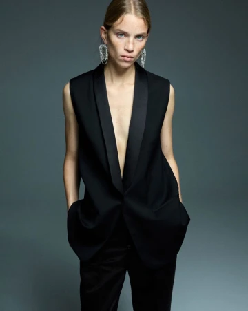 Fashion shot of a woman in a black sleeveless suit with statement earrings against a gray background in a studio setting.