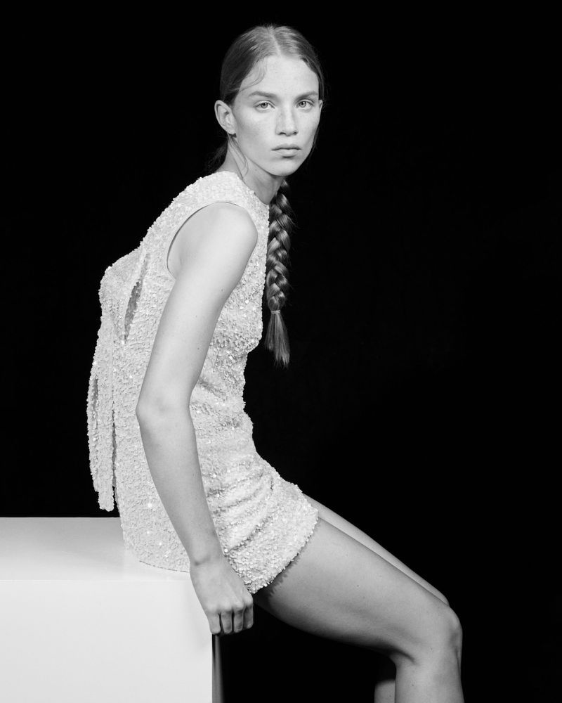 Monochrome portrait of a woman with a braid, seated in a sparkling mini-dress against a black backdrop, in a fashion-forward pose.